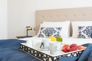 Un lit bien fait, avec un plateau contenant divers éléments pour le petit-déjeuner, notamment des fruits et des tasses, une lampe de chevet et des oreillers à motifs.