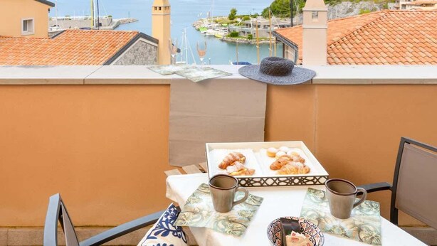 Ein Balkon mit Frühstückstisch, Hut und Blick auf das Meer und den Hafen.