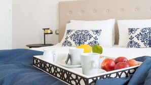 Ein ordentlich gemachtes Bett mit einem beige farbenen Kopfteil, weißen Kissen mit blauen Mustern und einem Tablett mit Früchten und Tassen auf einer blauen Bettdecke.