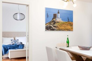 A room with a bed, a dining table, a painting of a lighthouse, and a hanging light fixture.