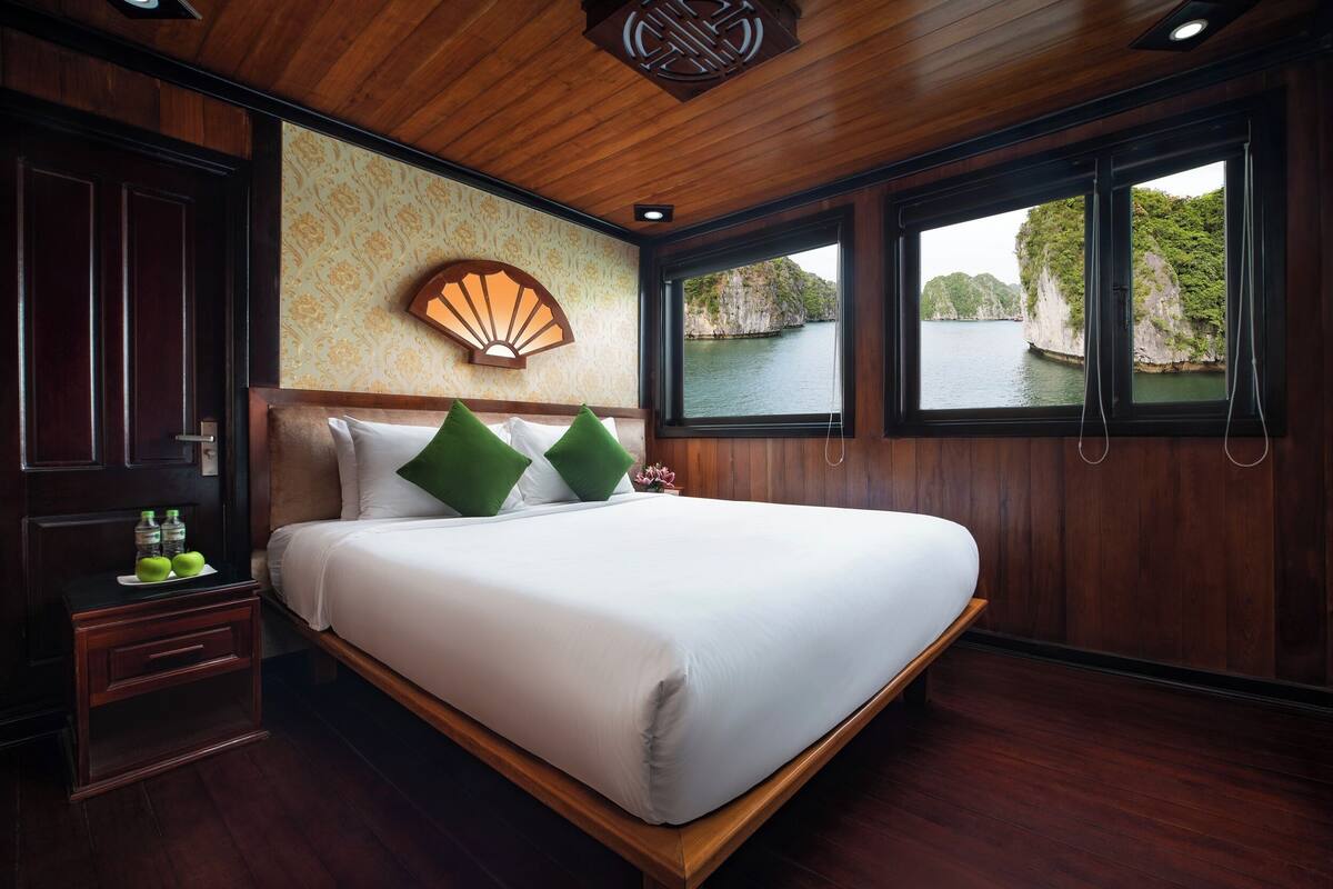 A wooden-paneled room with a bed, a nightstand, and a window with a view of a body of water and cliffs.