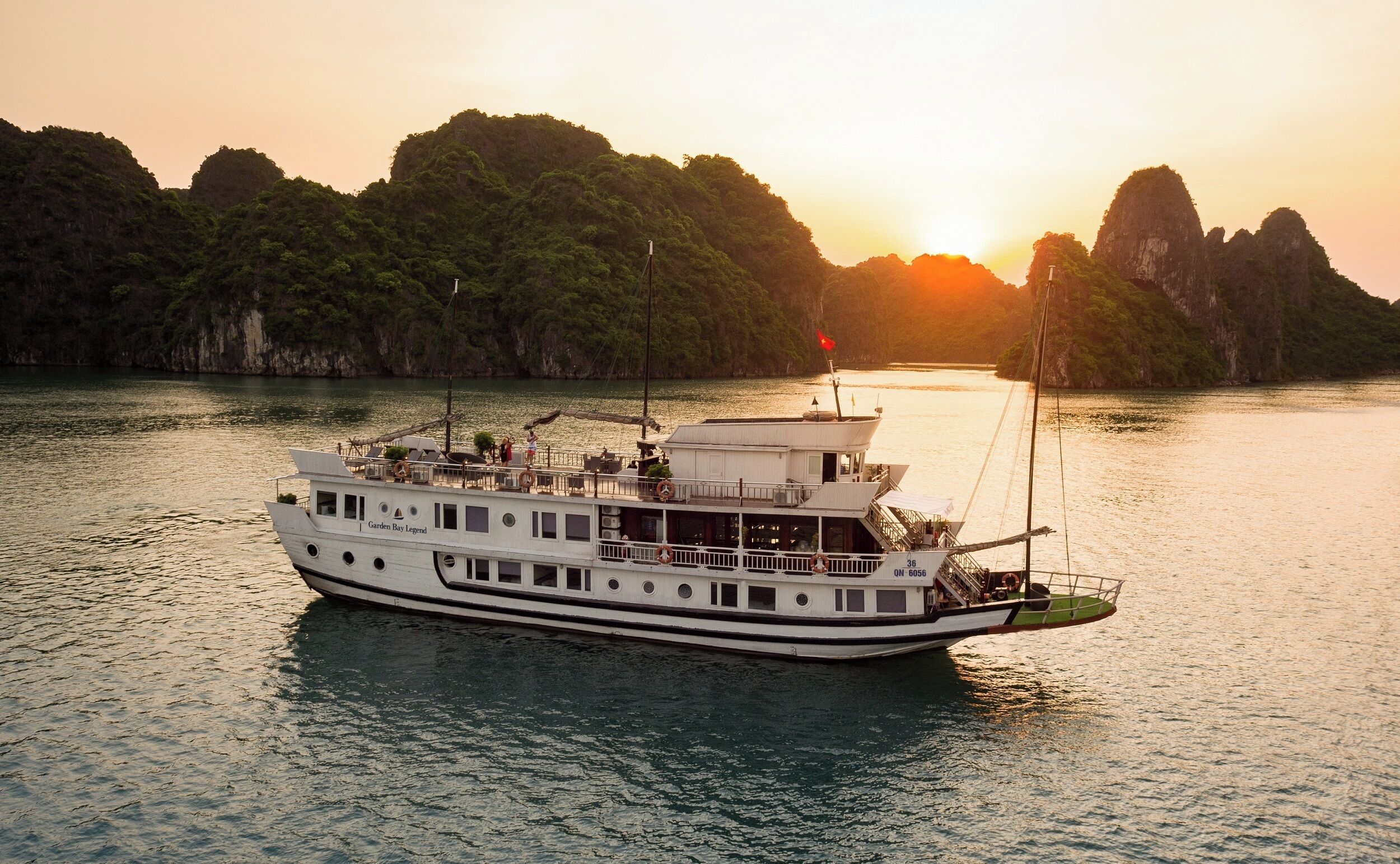 A large white boat named 'Garden Bay Legend' with multiple decks and a covered upper deck, sailing on calm waters with a backdrop of lush green hills and a setting sun.