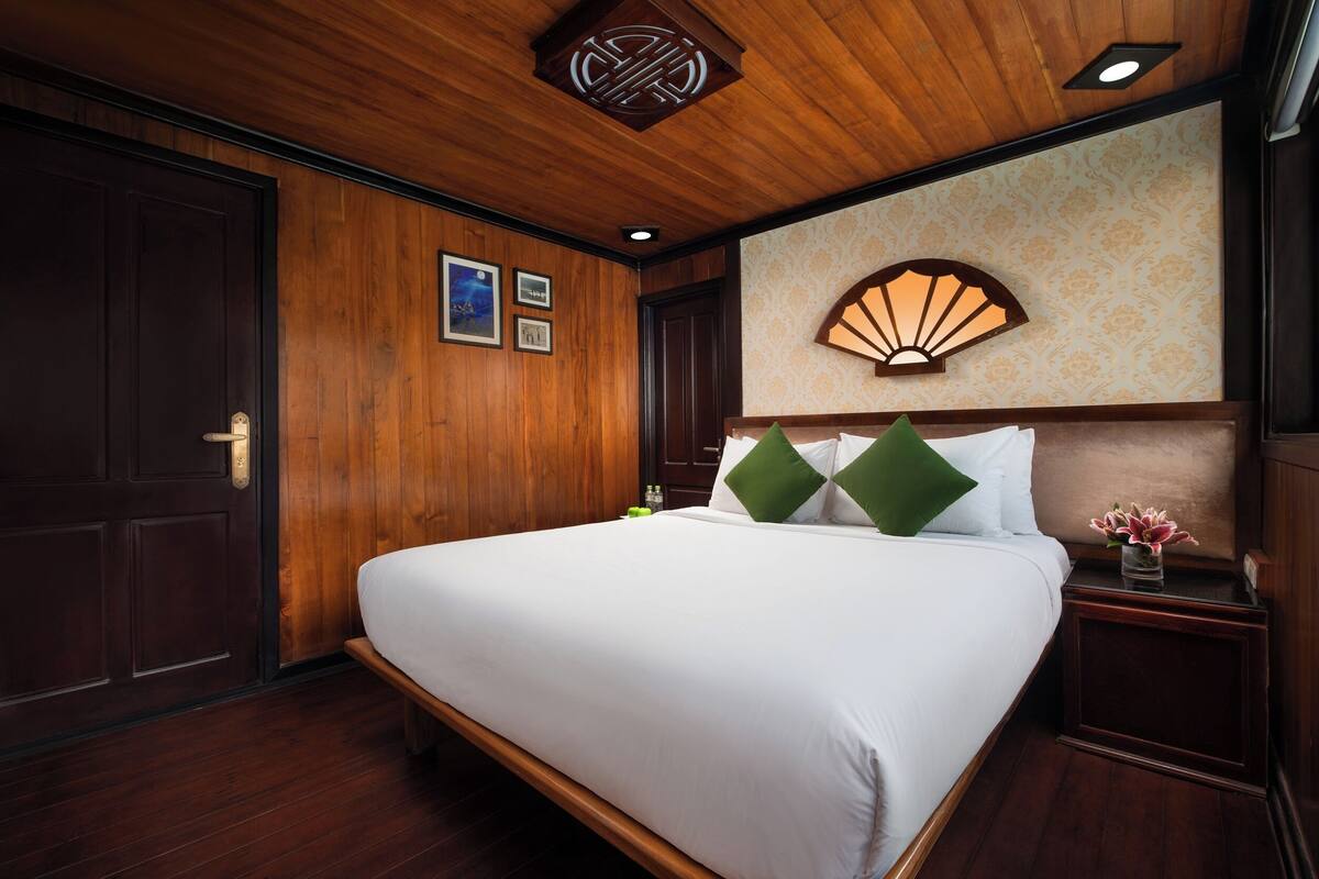 A hotel room with a wooden ceiling, a bed with white linens, green pillows, a nightstand with a vase of flowers, and a wall-mounted fan.