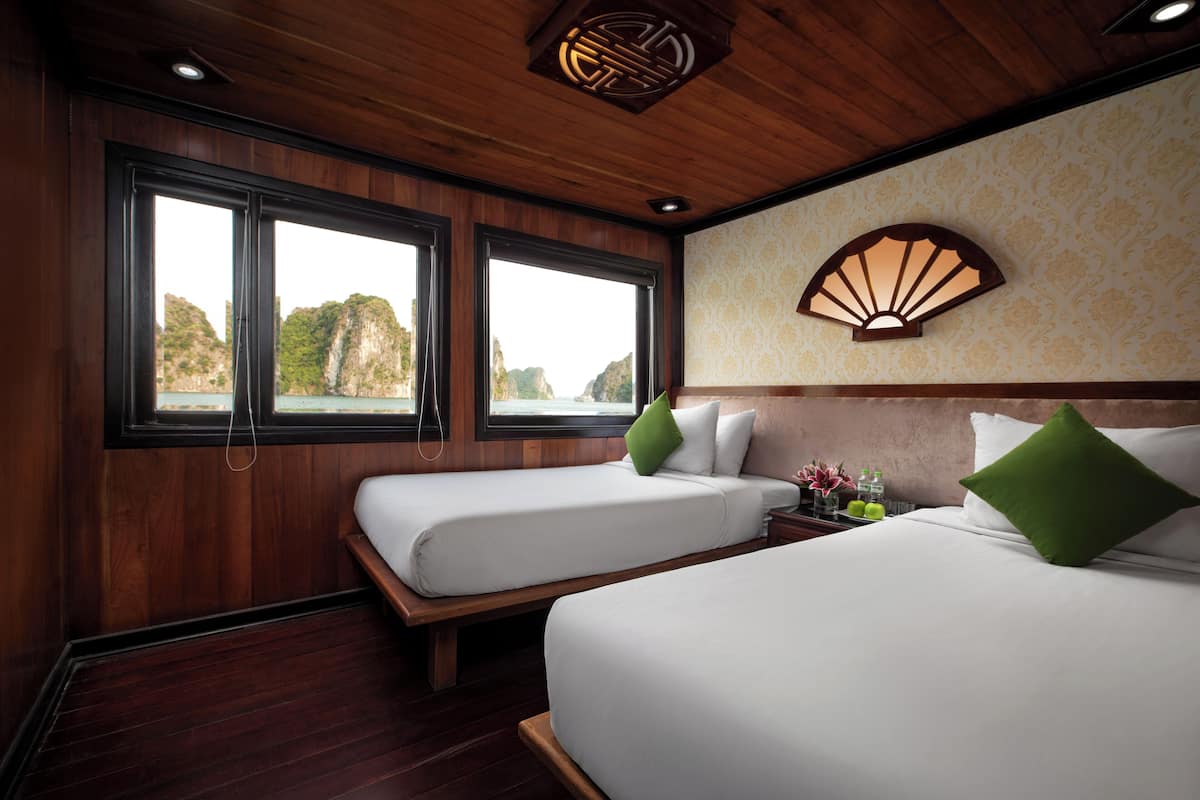 A hotel room with two beds, wooden flooring, large windows, a decorative wall piece, and a nightstand with a lamp and flowers.