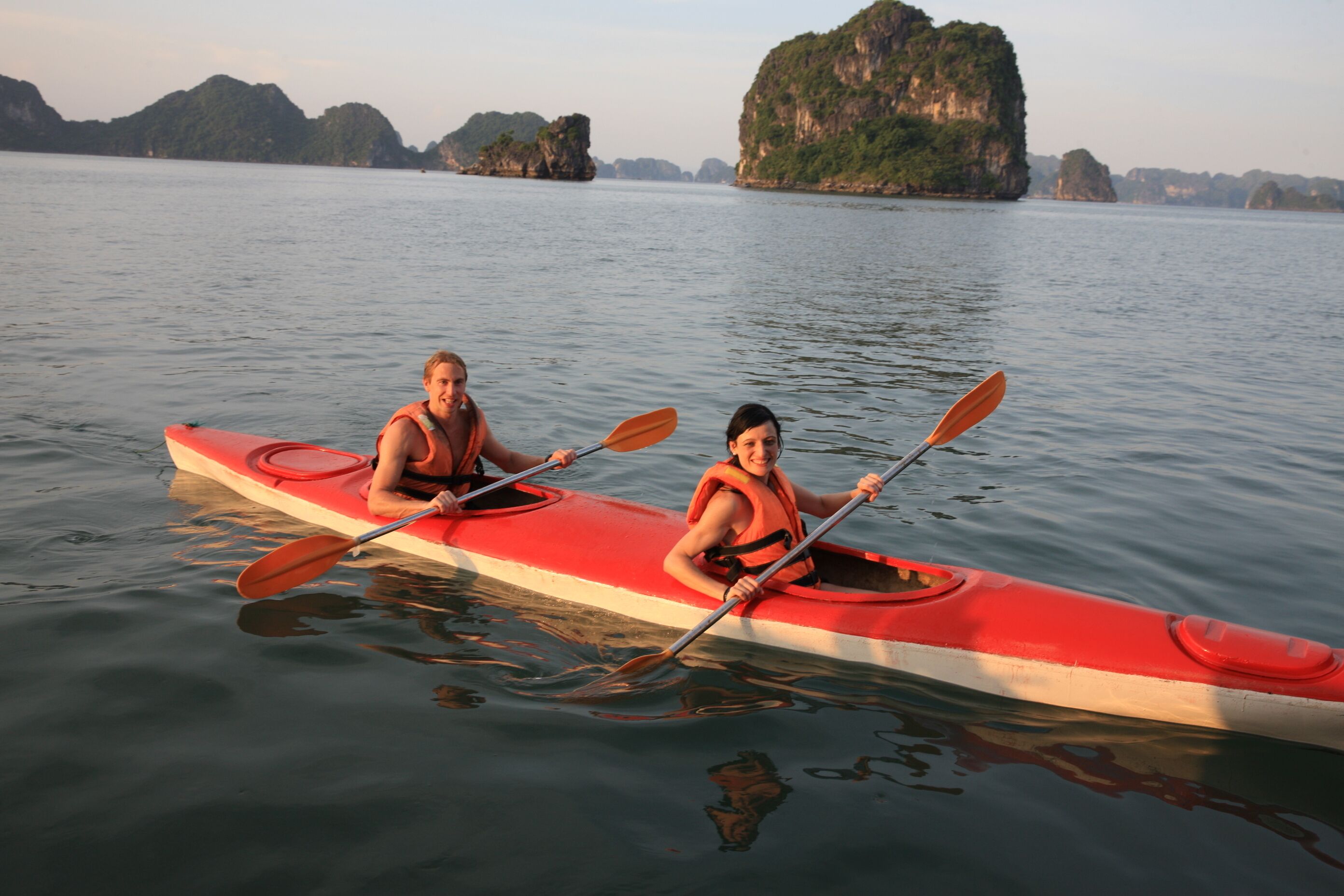 Two people kayaking on a calm body of water with a red kayak, surrounded by natural rock formations.