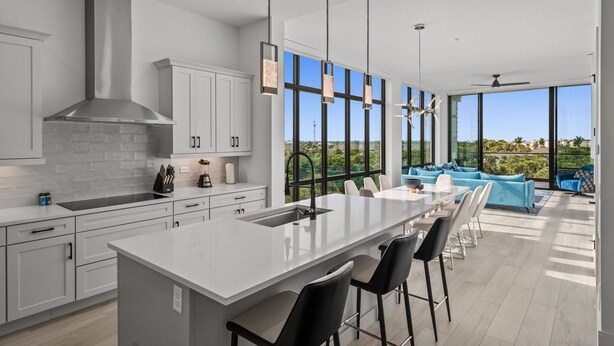 A modern kitchen with a large island, white cabinetry, and a view of the outdoors through large windows.
