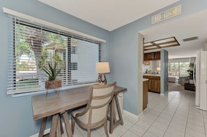 A modern kitchen with wooden cabinets, a dining area with a wooden table and chairs, and a view of the outside through large windows with blinds.