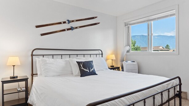 Una camera da letto con struttura letto in metallo, biancheria da letto bianca, un comodino con lampada, una finestra vista montagna e sci montati sulla parete.