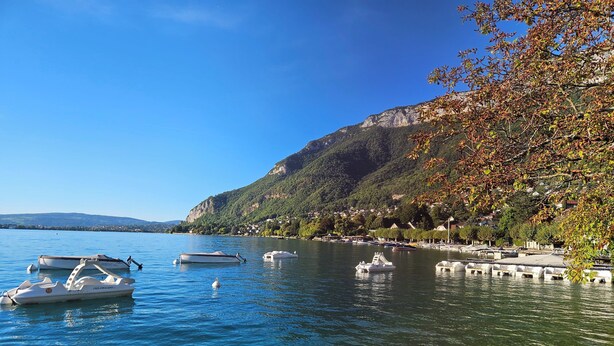 Ein ruhiger See mit Booten, klarem Wasser und einem Hang mit Bäumen und Häusern.