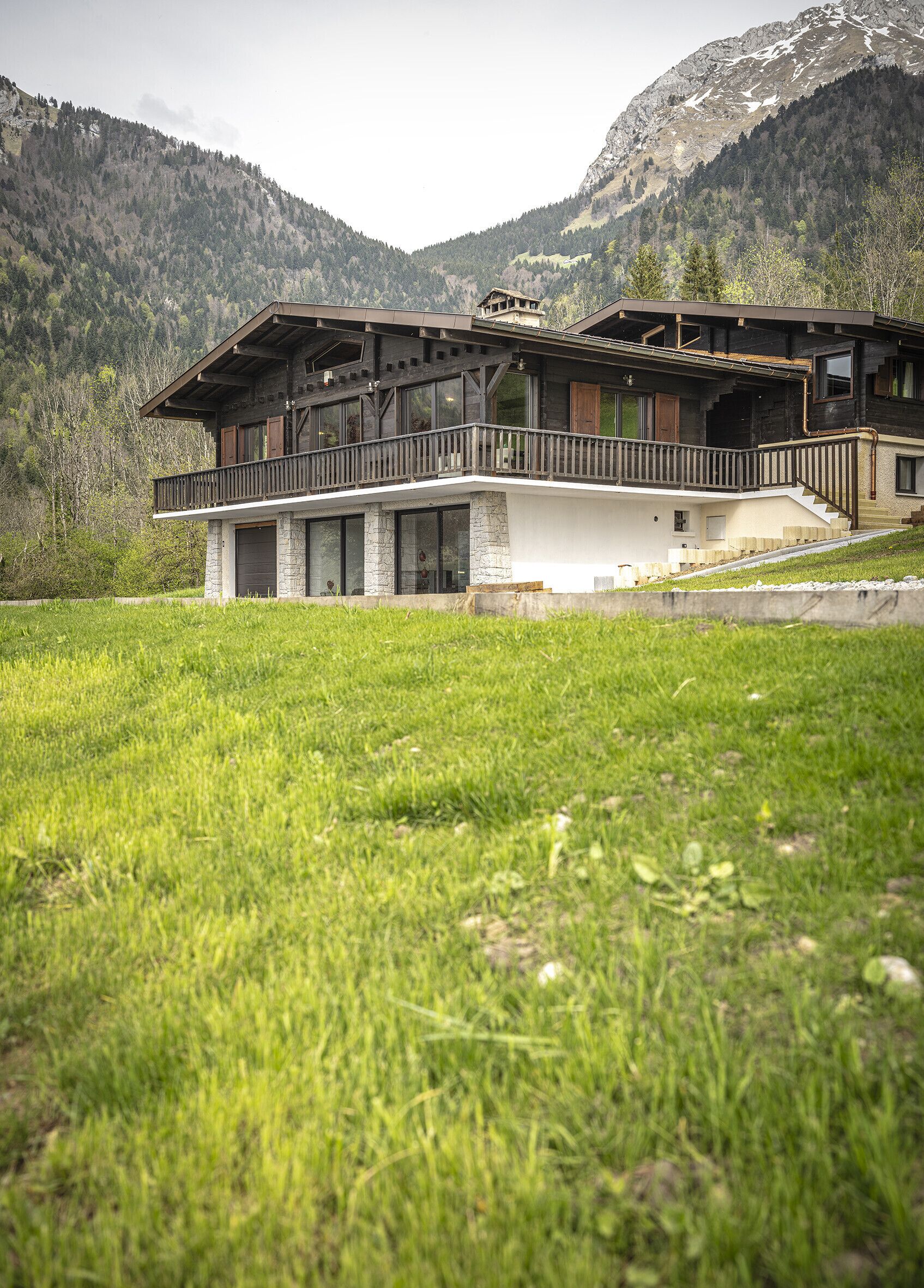 A two-story chalet with a balcony, stone foundation, and a view of a mountainous landscape.