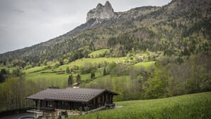 Eine rustikale Holzhütte mit geneigtem Dach, umgeben von üppiger Vegetation und einem Bergpanorama.