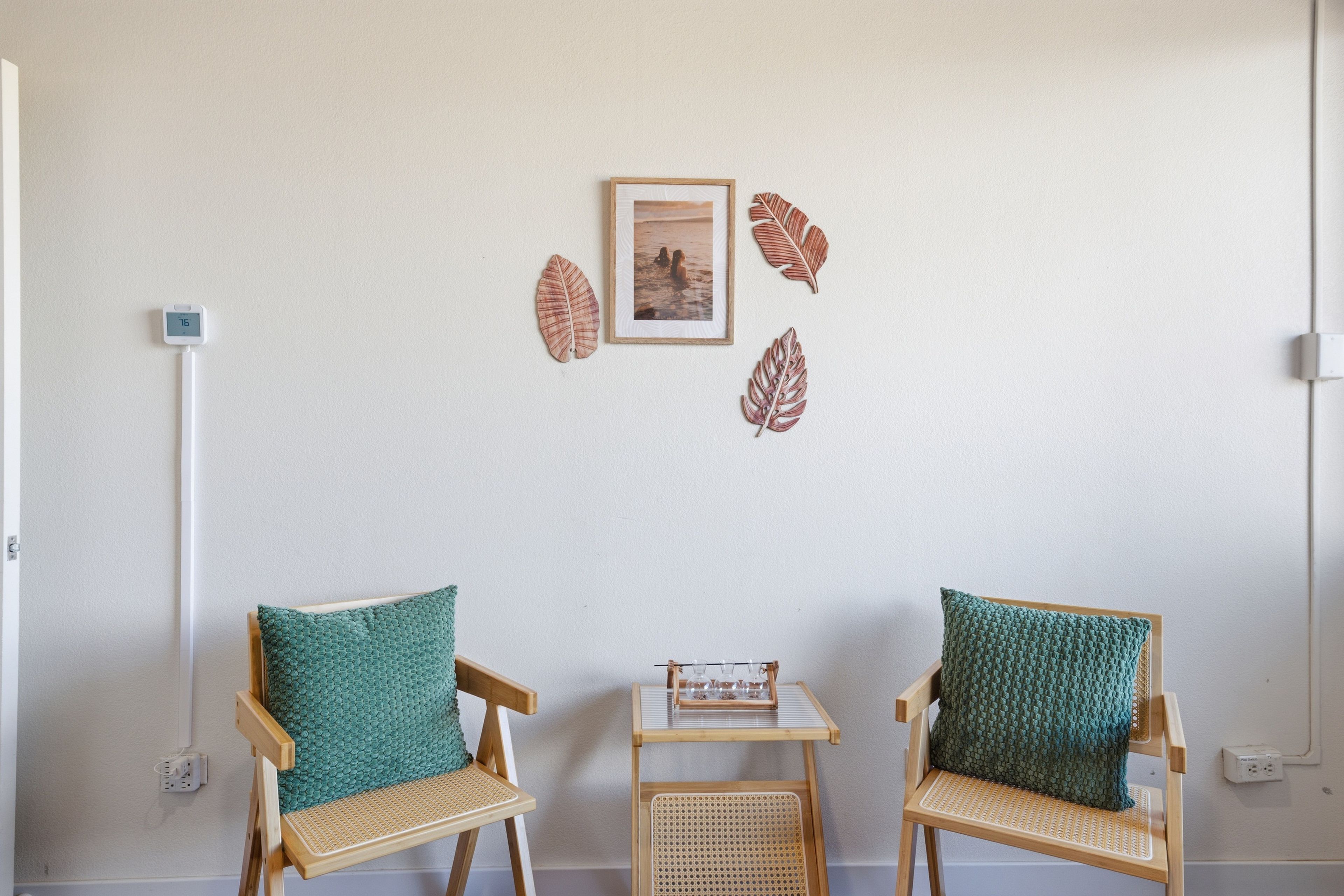 Une pièce comprenant deux chaises en bois avec des assises en osier et des coussins verts, une petite table sur laquelle se trouve un livre, et un tableau encadré accroché au mur.