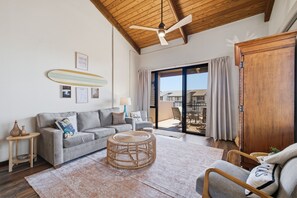A living room with a grey sofa, a wicker coffee table, a ceiling fan, and a view of a balcony with outdoor furniture.