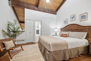 A bedroom with a wooden headboard, a bed with a beige bedspread, a nightstand with a lamp, a wooden chair, and a potted plant.