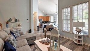 Ein geräumiges Wohnzimmer mit einer großen Eckcouch, einem Couchtisch und Blick auf die Küche und den Essbereich durch große Fenster.