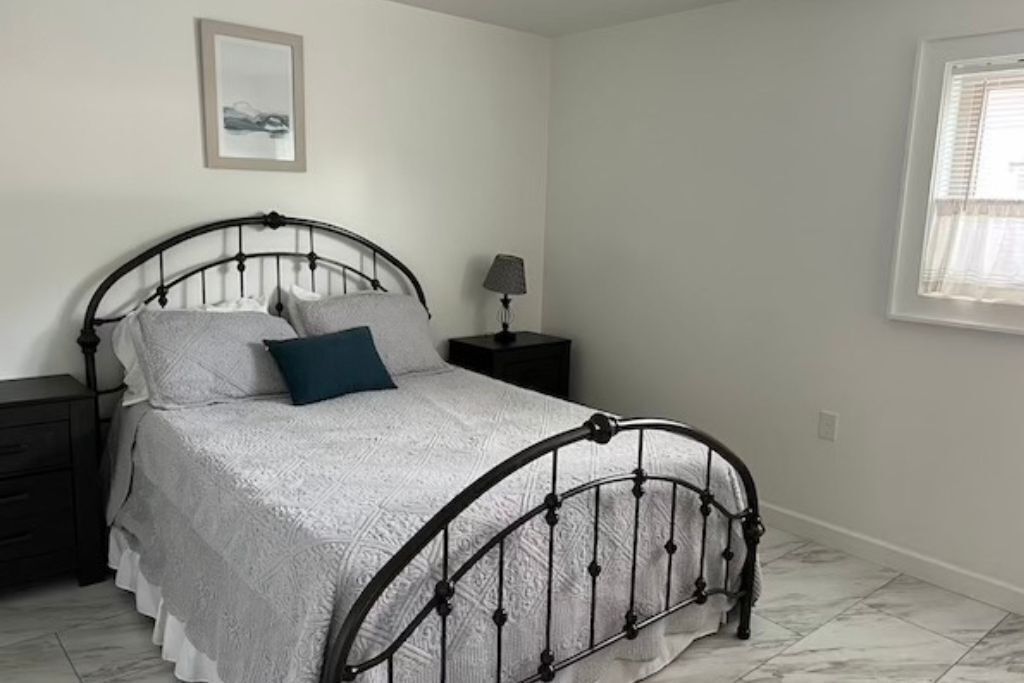 A bedroom with a metal bed frame, a white bedspread, a blue pillow, a bedside table with a lamp, and a window with blinds.