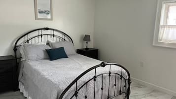 A bedroom with a metal bed frame, a white bedspread, a blue pillow, a bedside table with a lamp, and a window with blinds.
