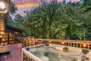 Un jacuzzi en una terraza de madera con sillas y una mesa, rodeado de árboles.