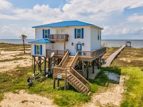 A two-story beach house with a blue roof, a deck, and a pier leading to the water.