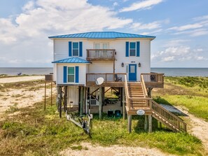 A two-story beach house with a blue roof, a balcony, and a deck with stairs leading to the entrance. - Dreamcatcher - 2814 Bienville 4 Bedroom Home by RedAwning (Dauphin Island)