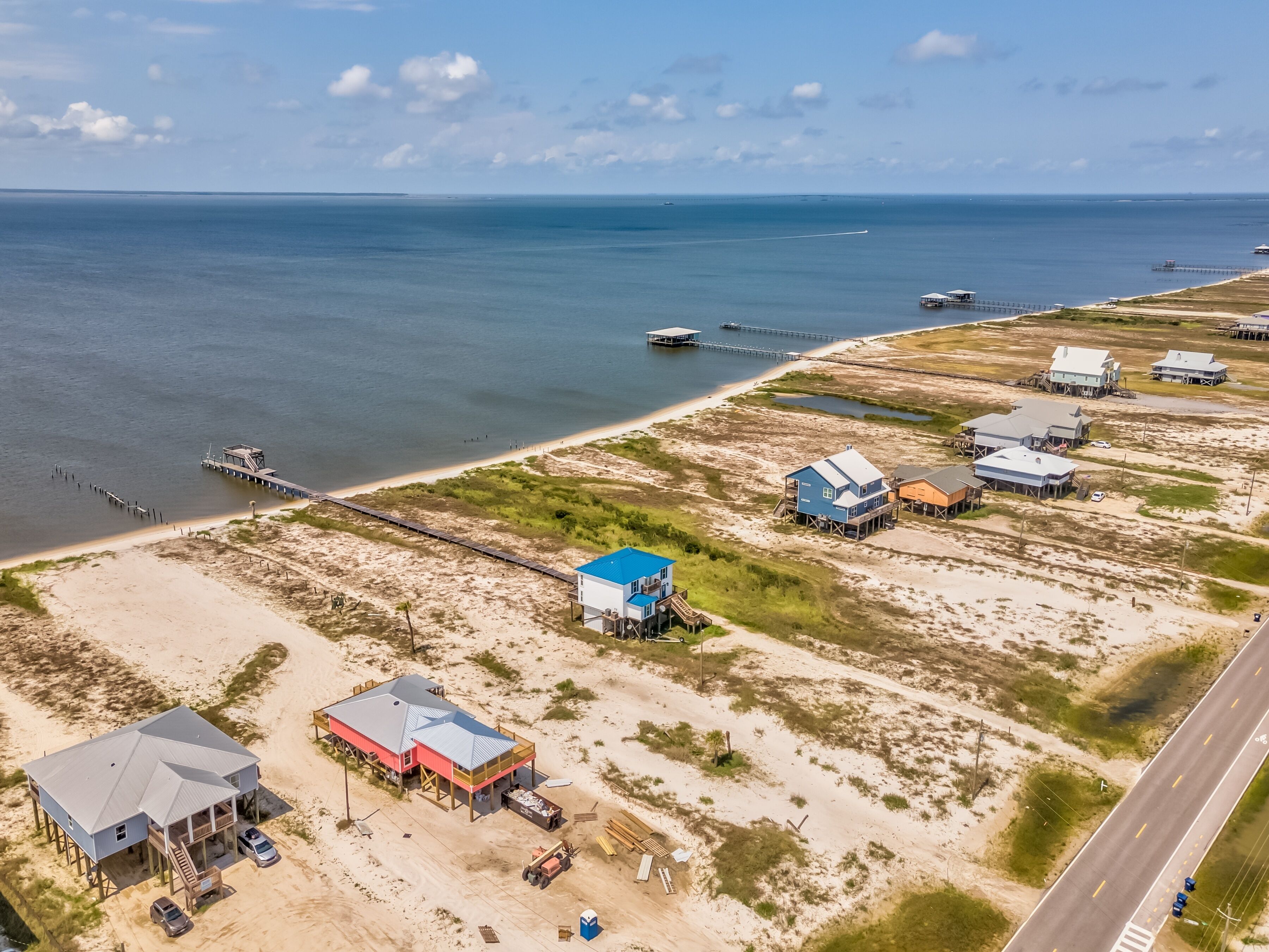An aerial view of a coastal area with a pier extending into the water, several buildings with elevated structures, and a road running parallel to the beach.