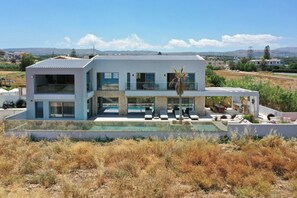 A modern two-story house with a swimming pool, large windows, and a view of the surrounding landscape. - Beachfront Villa with Eco heated Pool, Hot Tub, Playroom and walk to restaurants (Rethymno)