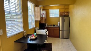 A compact kitchen with a stove, microwave, and refrigerator. There is a dining table with a basket of fruit and a bottle of water. - Scott Beach Villa #5 (St. Thomas)