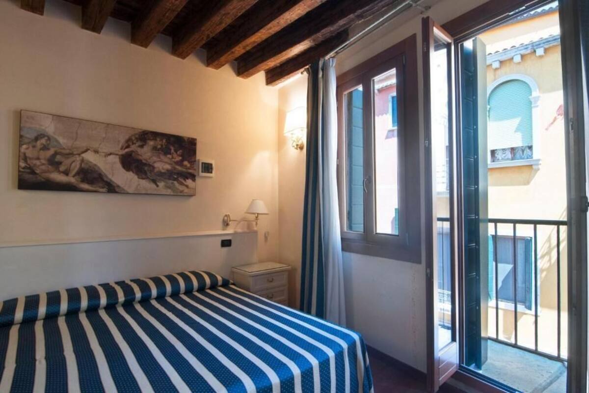 Una stanza con un letto, un quadro, un comodino, una lampada e una porta scorrevole in vetro che dà su una zona esterna.