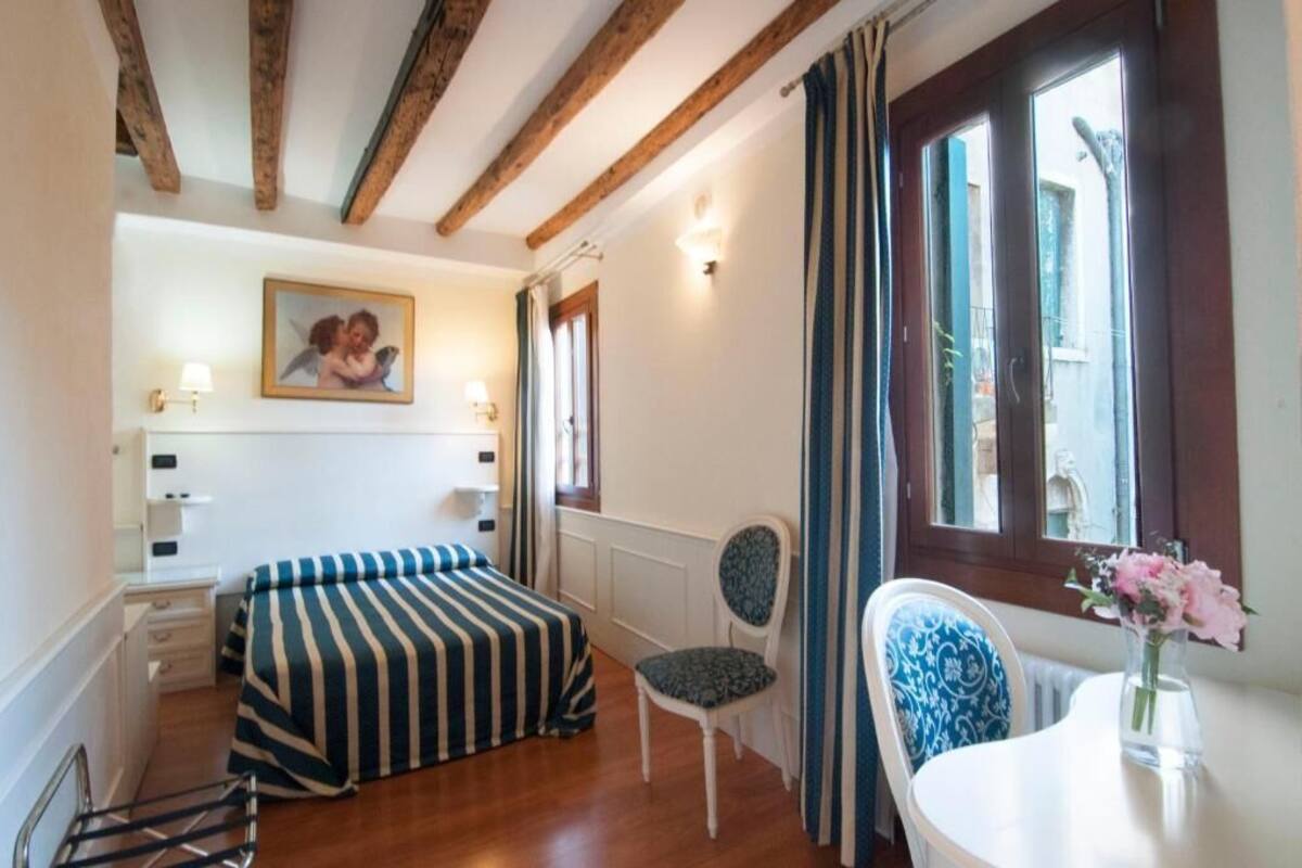 Una camera da letto con un letto, una sedia, un tavolo con un vaso di fiori, una finestra con tende e un quadro appeso al muro.