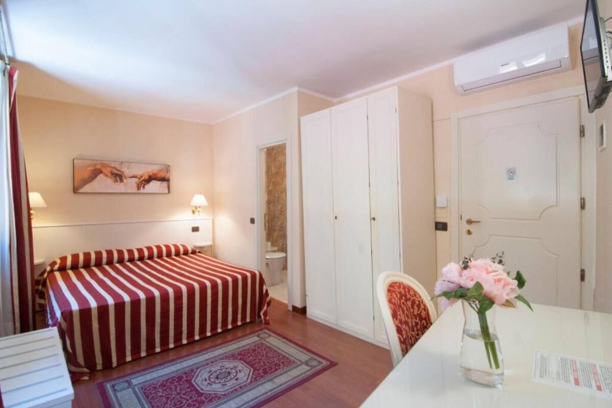 Una camera d'albergo con un letto, una scrivania, una sedia, un vaso con fiori e un quadro appeso al muro.