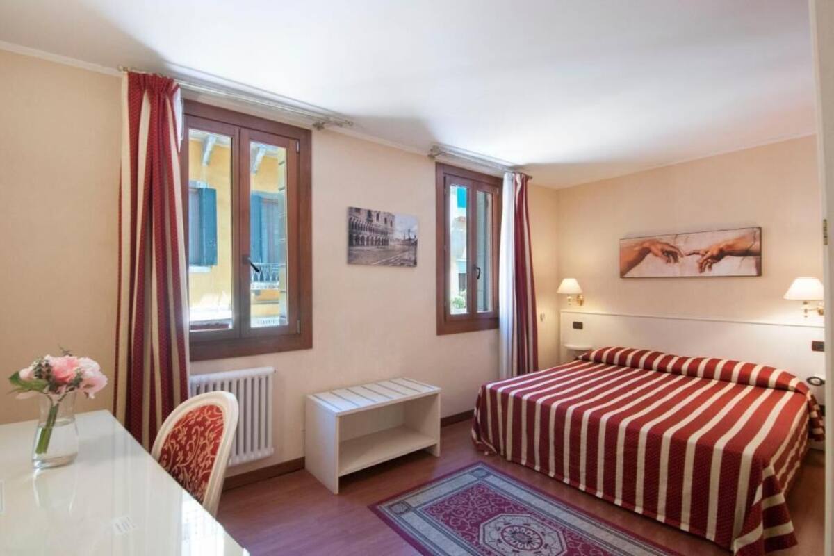 Una camera da letto con un letto, una scrivania, una sedia, un termosifone e due finestre con tende.