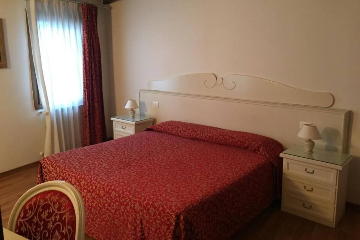 Una camera da letto con un letto, due comodini, una sedia, una finestra con tende e un termosifone.