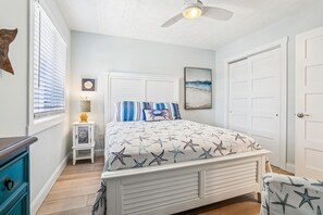 Une chambre à coucher avec un lit, un ventilateur de plafond, une table de chevet, une lampe, un tableau encadré et une porte.