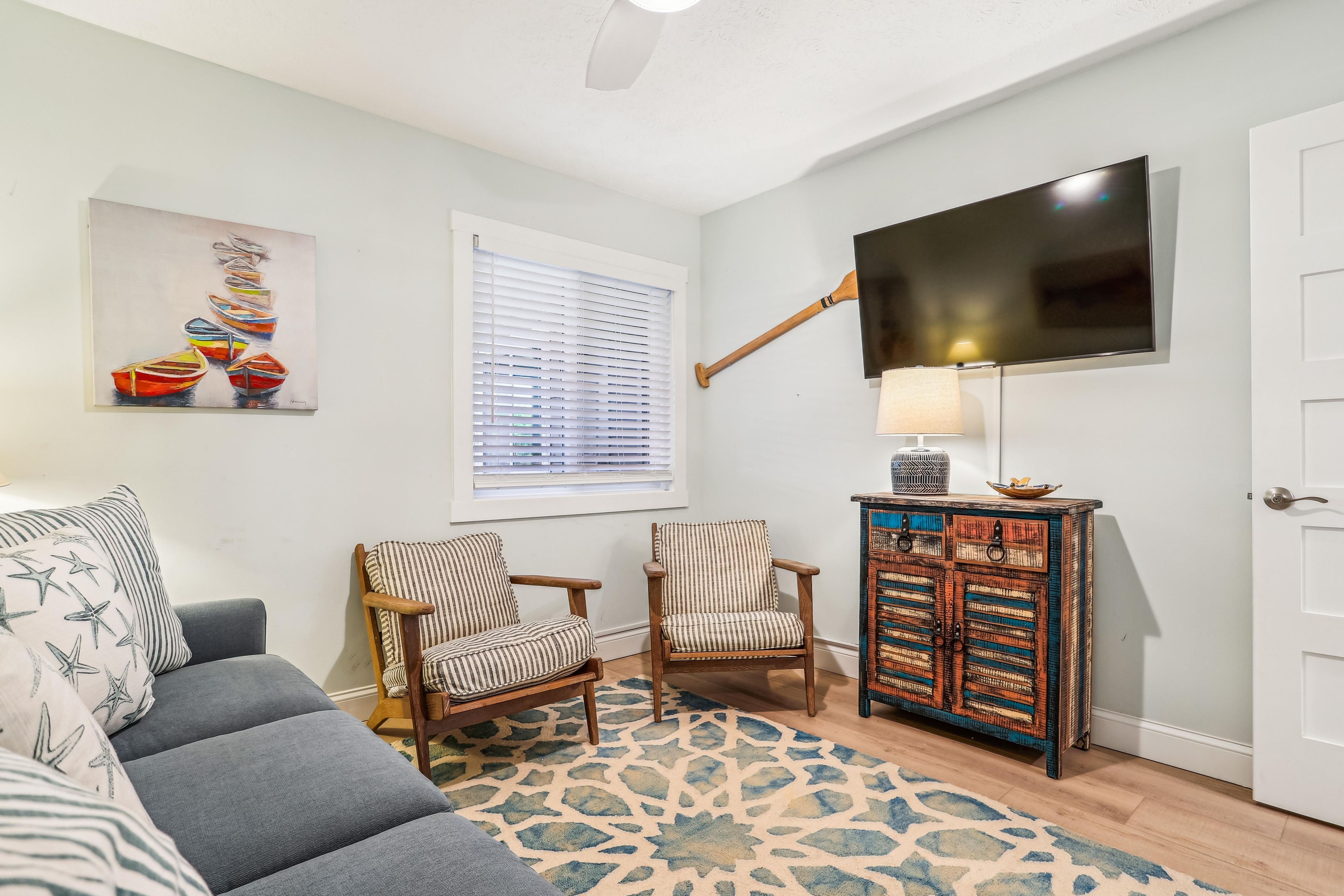 A living room with a grey sofa, two striped armchairs, a patterned rug, a television mounted on the wall, and a cabinet with decorative items.