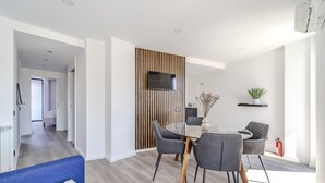 A modern living room with a dining area, featuring a TV mounted on a wooden panel wall, a round dining table with chairs, and a blue sofa.