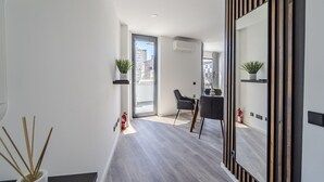 A modern, well-lit hallway with a wooden floor, a glass door leading to a balcony, and a dining area with a table and chairs.