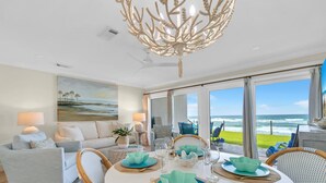 Un coin repas avec une table ronde dressée pour quatre personnes, entourée de chaises en osier. Une grande fenêtre offre une vue sur l’océan.