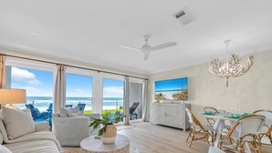 Un vaste espace de vie avec une grande fenêtre donnant sur la plage, une table à manger entourée de chaises en osier et un ventilateur de plafond.