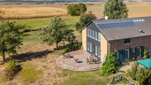 Une maison avec un toit équipé de panneaux solaires, un patio circulaire agrémenté de mobilier d’extérieur et une vue sur un champ de maïs.