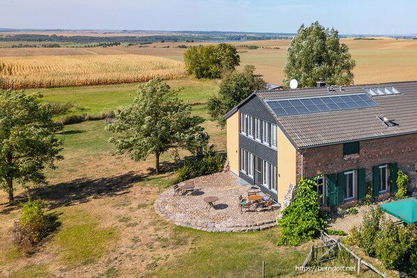 A house with a solar panel roof, a circular patio with outdoor furniture, and a view of a cornfield.