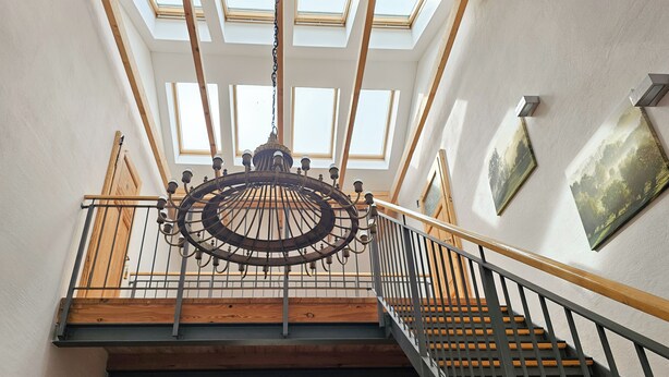 Un escalier avec une rampe en bois et des balustres noires, menant à un deuxième étage doté de lucarnes et d’un lustre.