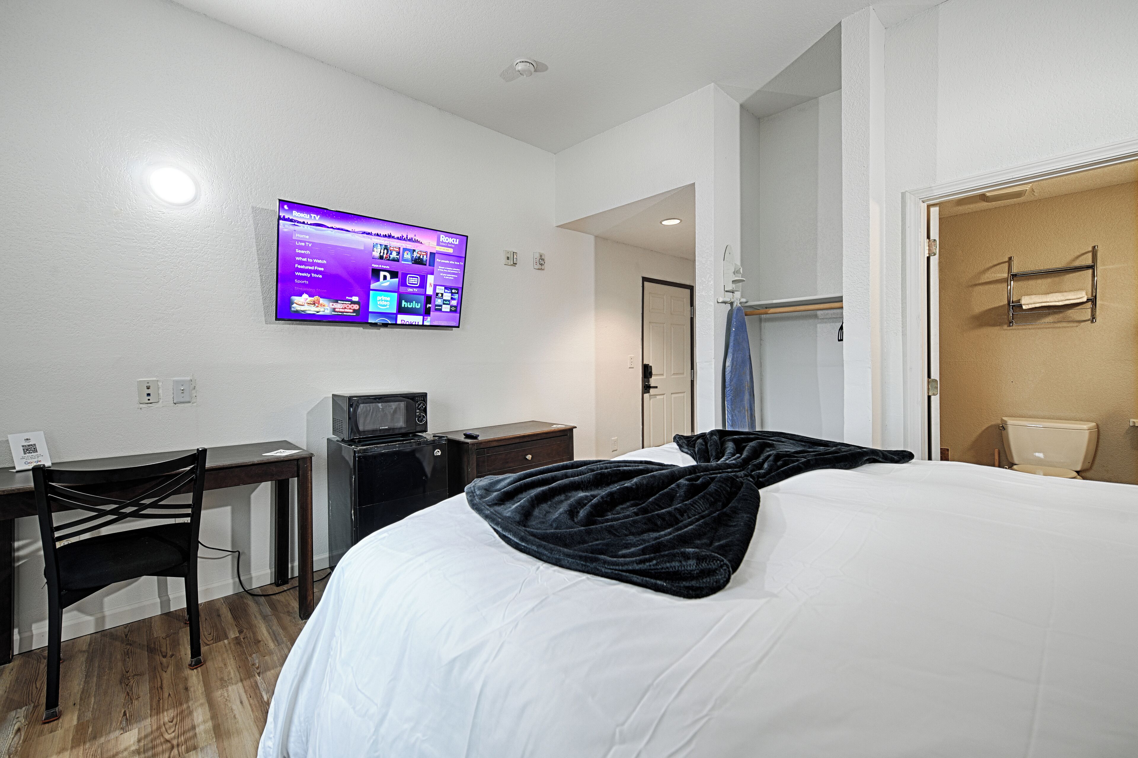 Une chambre d’hôtel avec un lit, un bureau équipé d’un ordinateur, un téléviseur fixé au mur et une salle de bain visible à travers une porte ouverte.