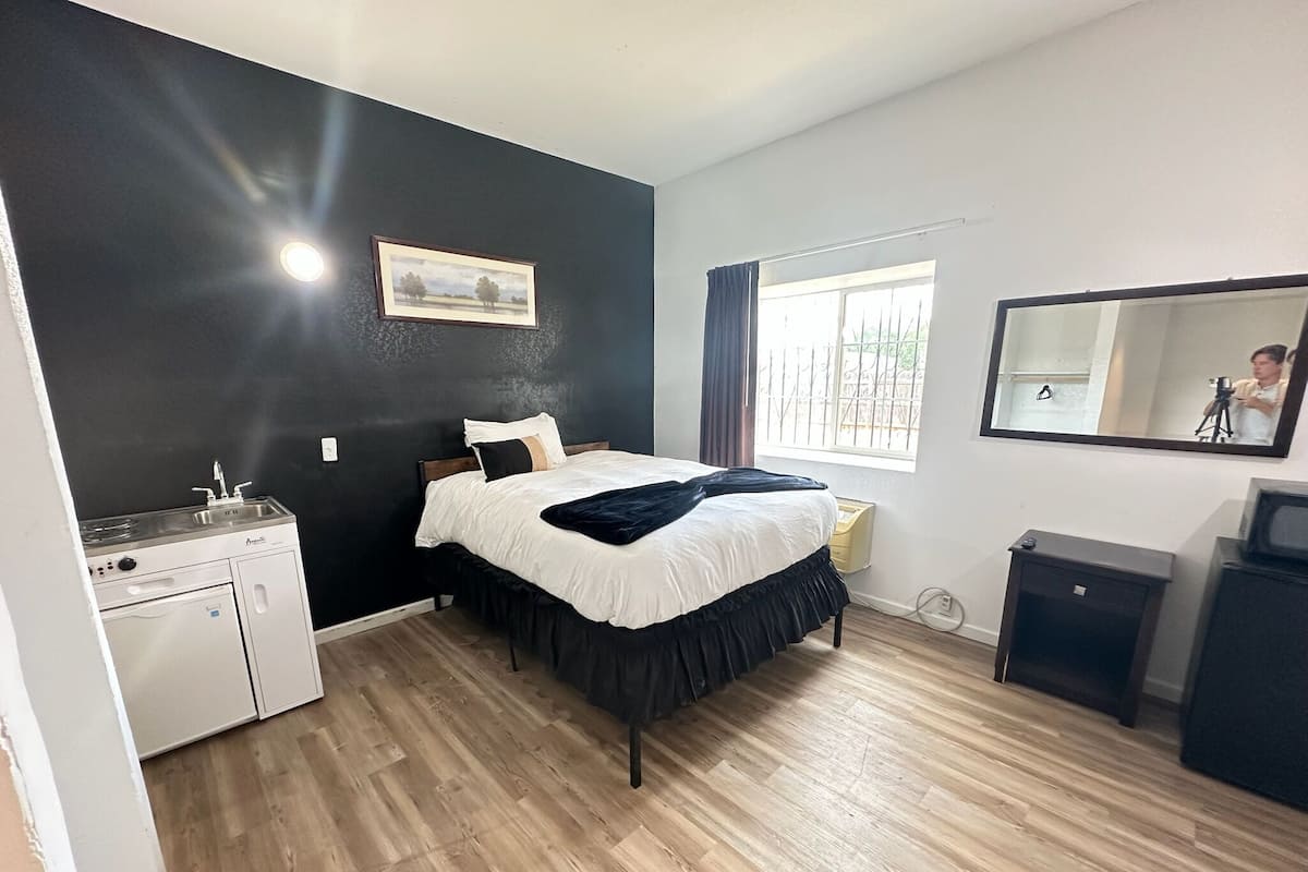A compact bedroom with a bed, a small kitchenette, a window with curtains, and a mirror on the wall.