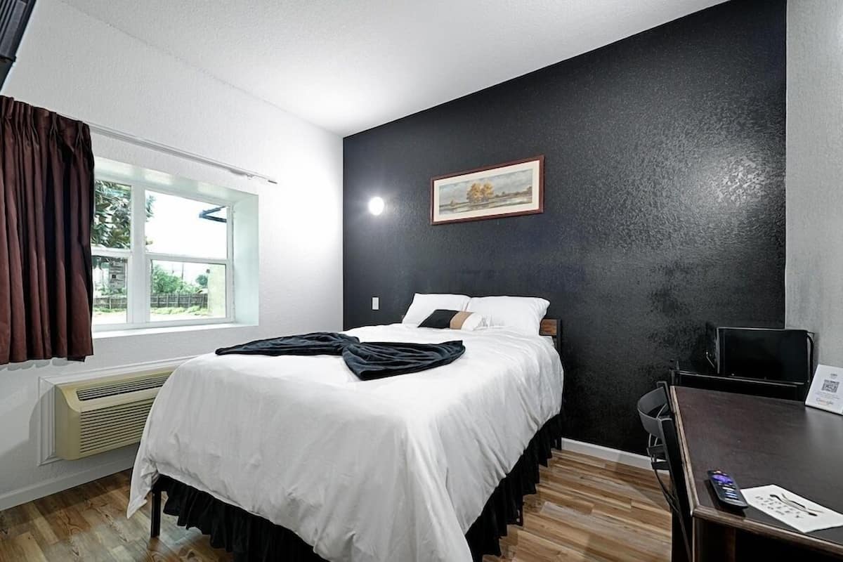 A hotel room with a bed, a desk, a chair, a window with curtains, a painting on the wall, and a heater.