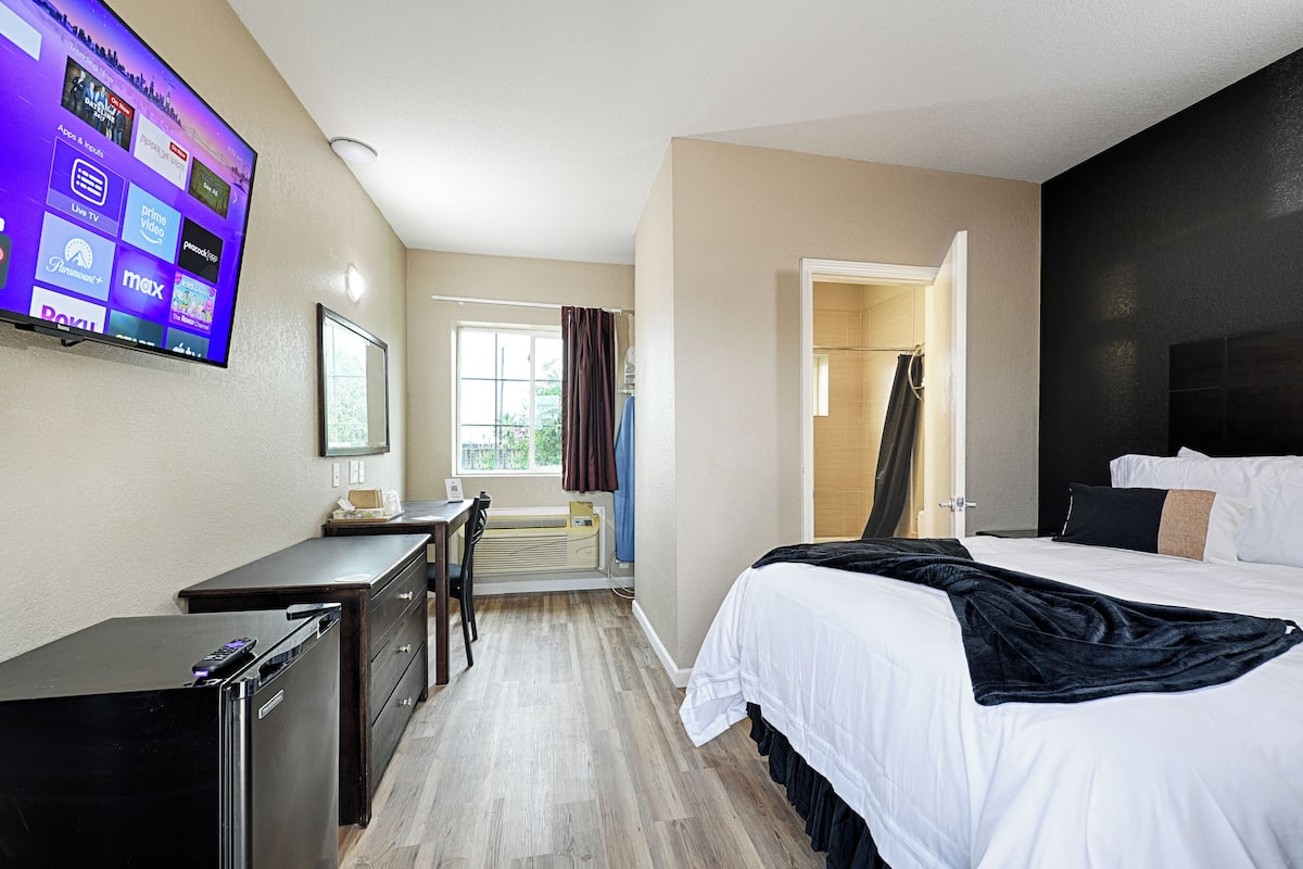 A hotel room with a bed, a desk, a TV mounted on the wall, and a window with curtains.