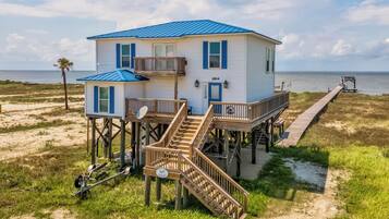 Une maison de plage à deux étages, avec un toit bleu, une terrasse et une jetée qui s’avance dans l’eau.