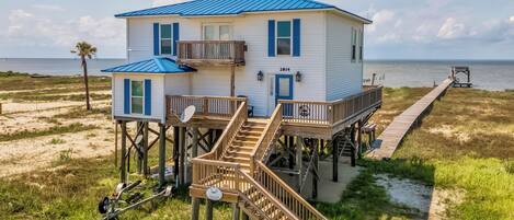 A two-story beach house with a blue roof, a deck, and a pier extending into the water.