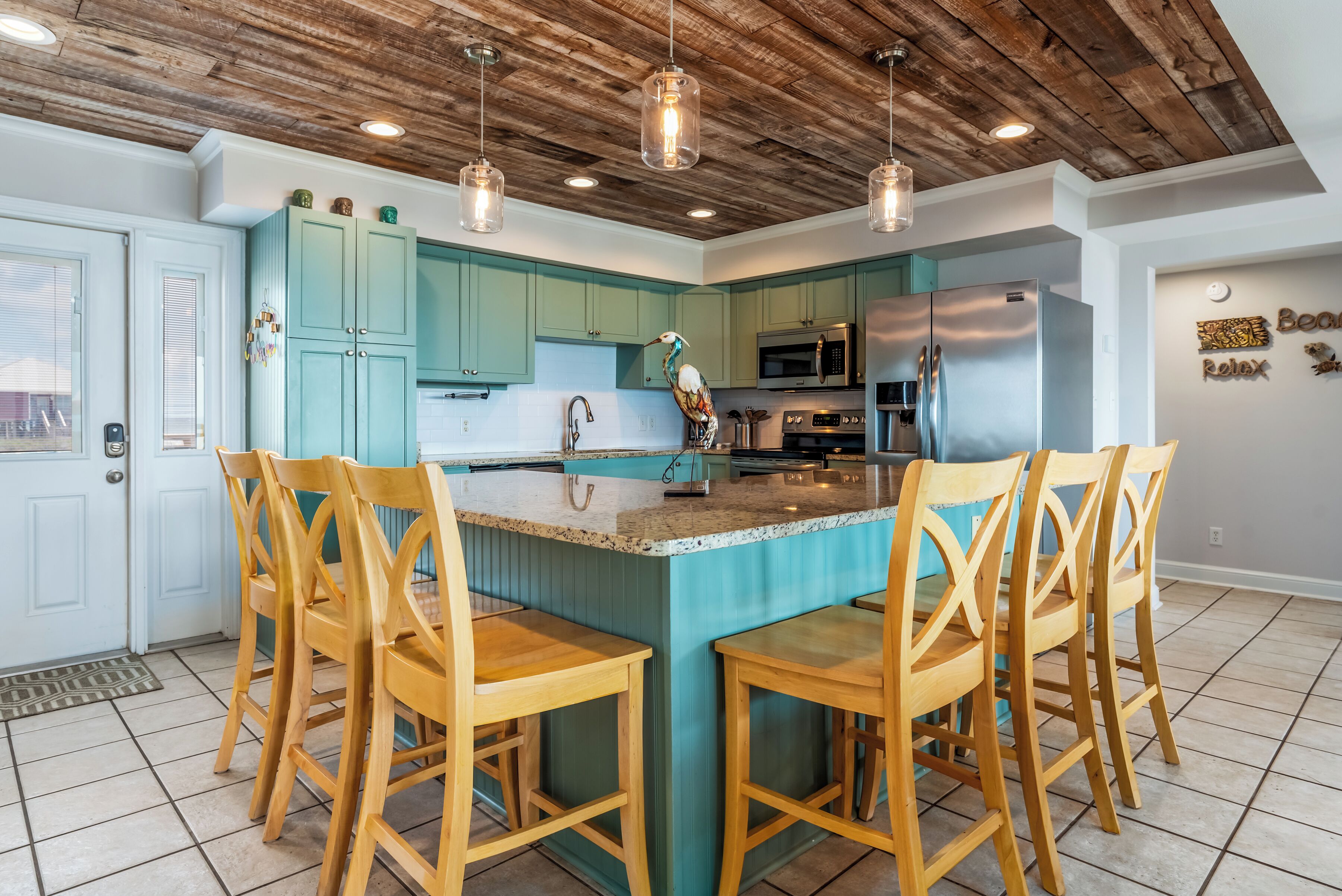 Une cuisine avec des meubles de couleur turquoise, un îlot central doté de tabourets de bar, un plafond en bois et des luminaires suspendus.