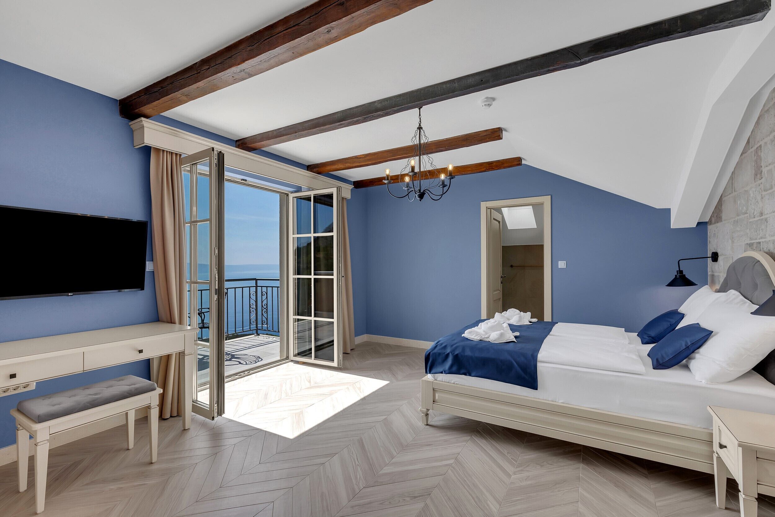 A bedroom with a bed, a desk, a bench, a TV, and a balcony with a view.