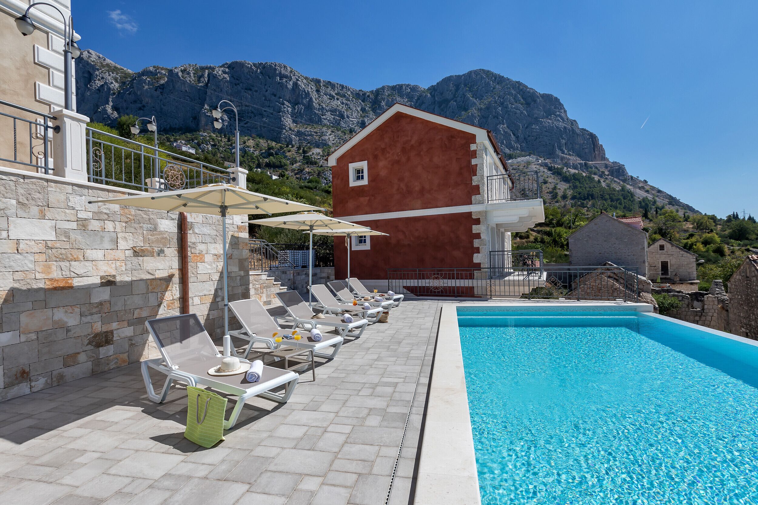 En swimmingpool med liggestole og parasoller, en stenvæg og et bjerg i baggrunden.